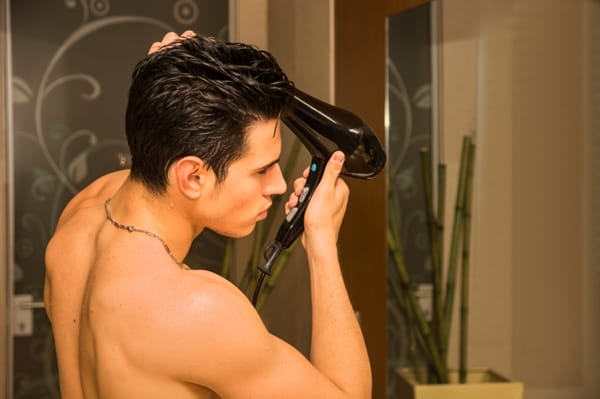 Shirtless young man drying hair with hairdryer, looking at camera at home